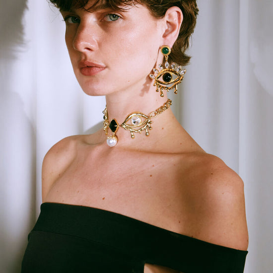 Woman wearing gold earrings and a necklace with a black top against a white curtain background