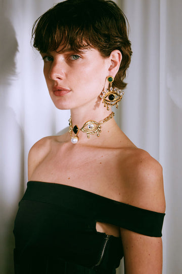 Woman wearing gold earrings and a necklace with a black top against a white curtain background